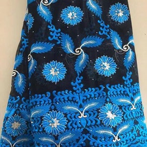May include: A folded piece of fabric with a black base and intricate blue floral embroidery. The design features large, stylized flowers and leaf patterns, with white accents. The edges are finished with a scalloped border in a vibrant blue.