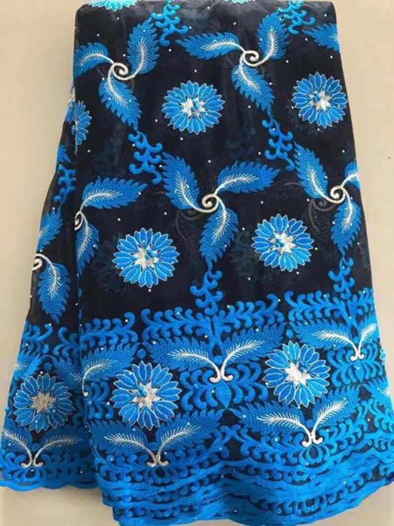 May include: A folded piece of fabric with a black base and intricate blue floral embroidery. The design features large, stylized flowers and leaf patterns, with white accents. The edges are finished with a scalloped border in a vibrant blue.