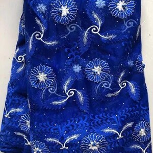 May include: Royal blue lace fabric with intricate white floral and leaf embroidery. The fabric features a detailed pattern of flowers, leaves, and swirling designs, creating a textured and elegant appearance. This fabric is suitable for dresses and other apparel.