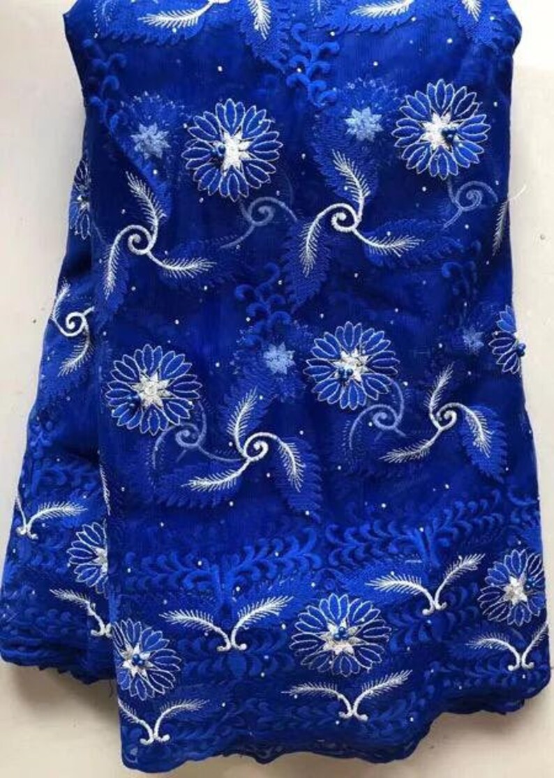 May include: Royal blue lace fabric with intricate white floral and leaf embroidery. The fabric features a detailed pattern of flowers, leaves, and swirling designs, creating a textured and elegant appearance. This fabric is suitable for dresses and other apparel.