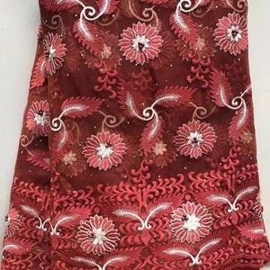 May include: Burgundy lace fabric with a floral and leaf pattern. The design features embroidered flowers and leaves in shades of red and white. The fabric is semi-sheer, with a delicate texture, suitable for apparel or craft projects.
