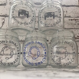 May include: Four glass jars with lids, each decorated with a different gaming-themed design. The designs include a video game controller, a headset with a microphone, and the text "Gaming Snacks".