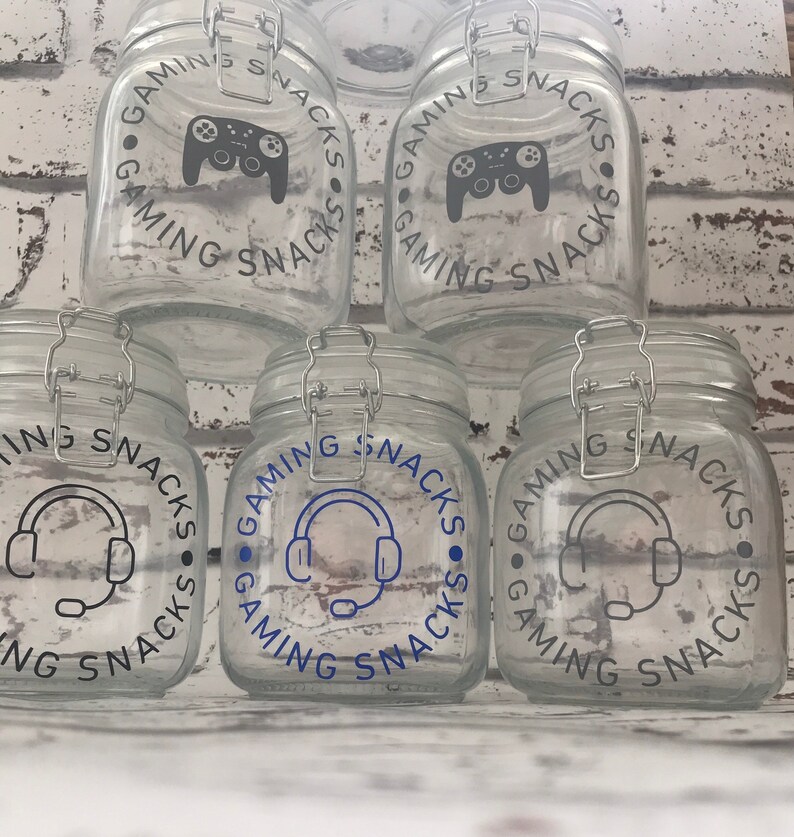 May include: Four glass jars with lids, each decorated with a different gaming-themed design. The designs include a video game controller, a headset with a microphone, and the text "Gaming Snacks".
