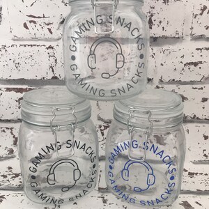 May include: Three glass jars with airtight lids. Each jar has a black and white graphic of a headset with the text "Gaming Snacks" around the graphic. The top jar has gray text, the middle jar has blue text, and the bottom jar has blue text.