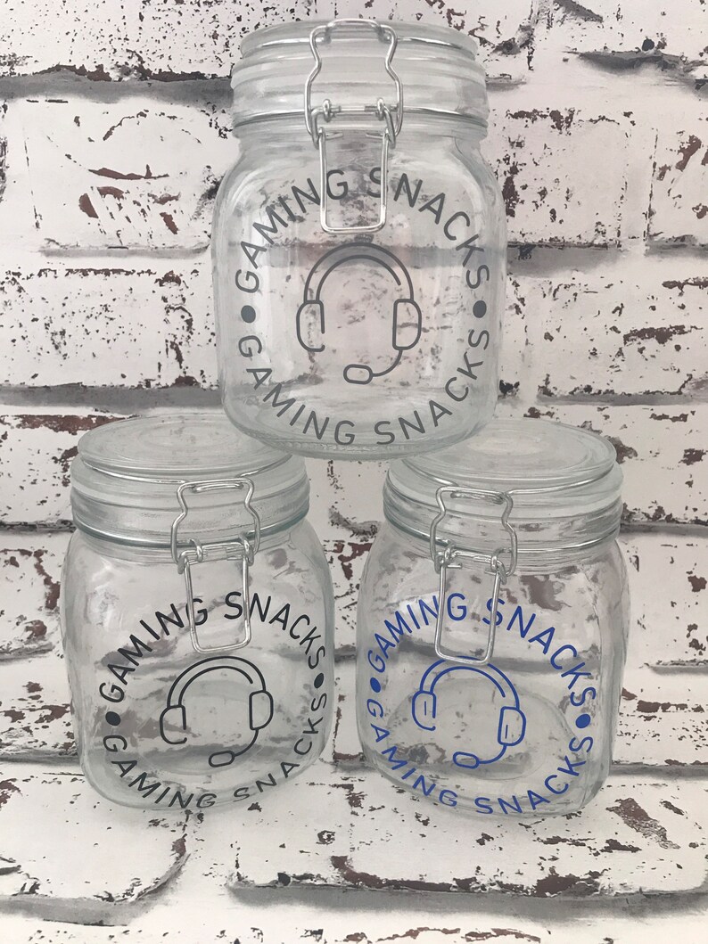 May include: Three glass jars with airtight lids. Each jar has a black and white graphic of a headset with the text "Gaming Snacks" around the graphic. The top jar has gray text, the middle jar has blue text, and the bottom jar has blue text.