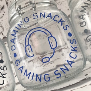 May include: A clear glass jar with a blue and white label that says "Gaming Snacks" with a headset icon. The jar has a silver lid with a latch.