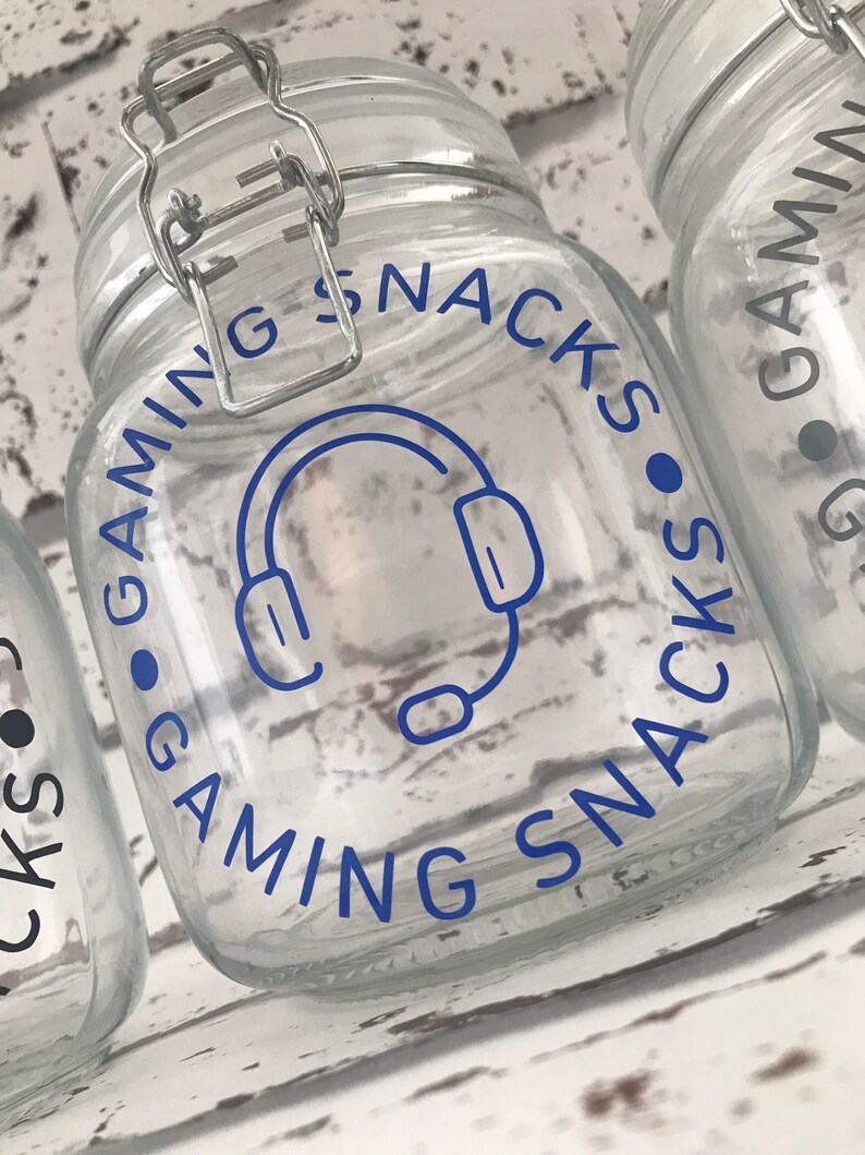May include: A clear glass jar with a blue and white label that says "Gaming Snacks" with a headset icon. The jar has a silver lid with a latch.