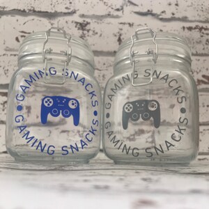 May include: Two clear glass jars with airtight lids. The jars are decorated with a blue and gray video game controller graphic and the text "Gaming Snacks".