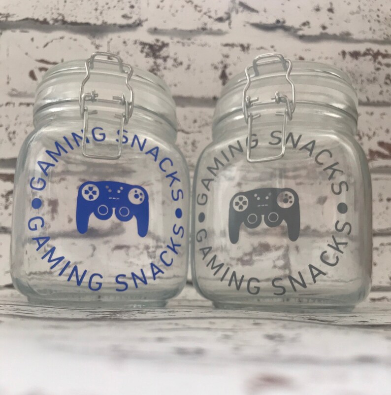 May include: Two clear glass jars with airtight lids. The jars are decorated with a blue and gray video game controller graphic and the text "Gaming Snacks".