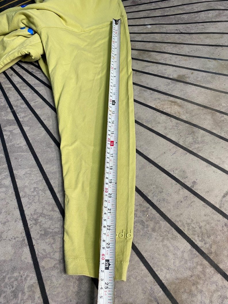 May include: A lime green long-sleeve shirt featuring the word "did" embroidered in a matching thread near the cuff. A white measuring tape is placed along the sleeve, indicating a length of approximately 24.5 inches from the shoulder seam.