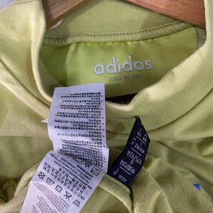 May include: A light lime green Adidas Fear of God t-shirt with a mock neck collar. The interior neck label displays the Adidas Fear of God logo. Attached care tags feature detailed washing instructions in multiple languages and size information.