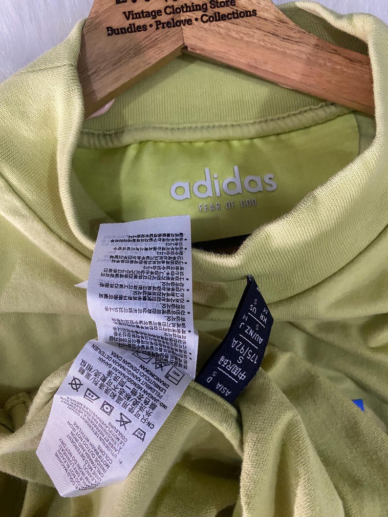 May include: A light lime green Adidas Fear of God t-shirt with a mock neck collar. The interior neck label displays the Adidas Fear of God logo. Attached care tags feature detailed washing instructions in multiple languages and size information.