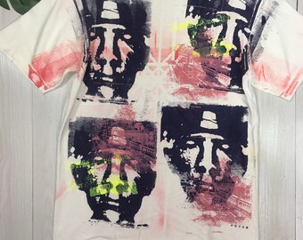 May include: White t-shirt with a multi-colored all-over print of a man's face with a hat. The print is in shades of blue, pink, red, and yellow. The shirt has a crew neck and short sleeves.