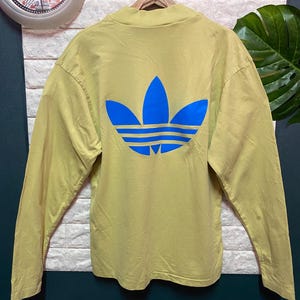 May include: A long-sleeve mock neck shirt in a muted olive green color. The back of the shirt features a large, bold, bright blue Adidas trefoil logo printed centrally. The garment is displayed on a wooden hanger against a textured wall.