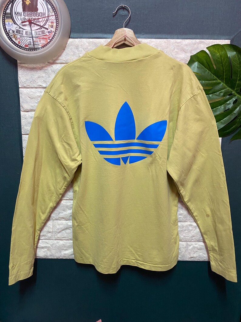 May include: A long-sleeve mock neck shirt in a muted olive green color. The back of the shirt features a large, bold, bright blue Adidas trefoil logo printed centrally. The garment is displayed on a wooden hanger against a textured wall.