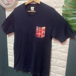 May include: Navy blue t-shirt with a pocket featuring a red, yellow, and white plaid pattern. The shirt has short sleeves and a crew neck. The pocket is rectangular and sewn onto the left chest area.