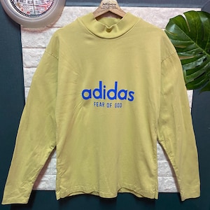 May include: A long-sleeve mock neck shirt in a muted yellow color. The front features the blue text "adidas" above the blue text "FEAR OF GOD". The shirt is displayed on a wooden hanger against a textured white and dark gray background.