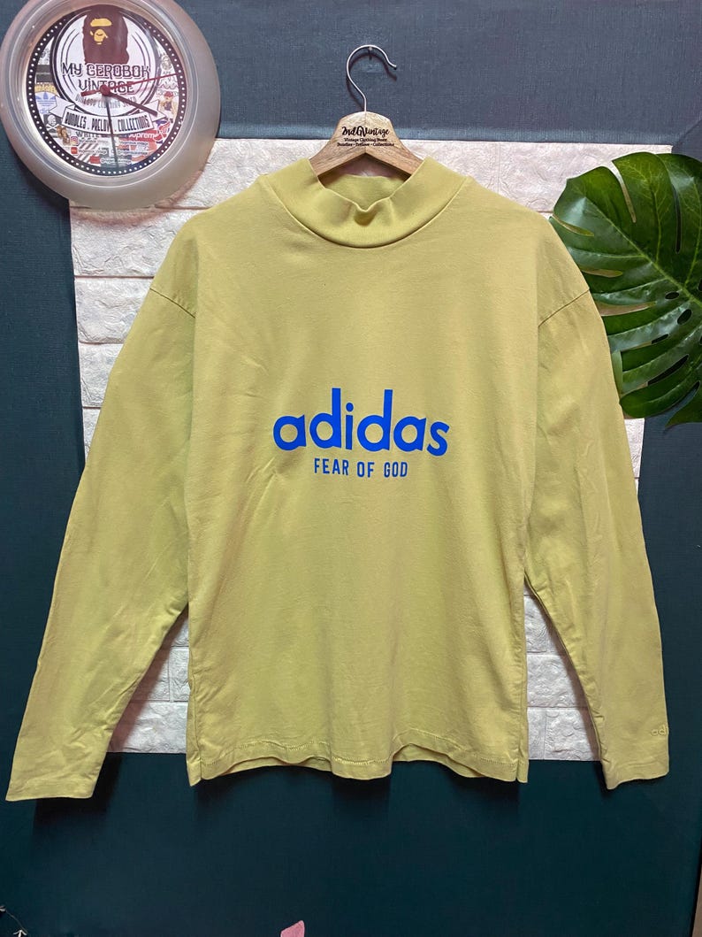 May include: A long-sleeve mock neck shirt in a muted yellow color. The front features the blue text "adidas" above the blue text "FEAR OF GOD". The shirt is displayed on a wooden hanger against a textured white and dark gray background.