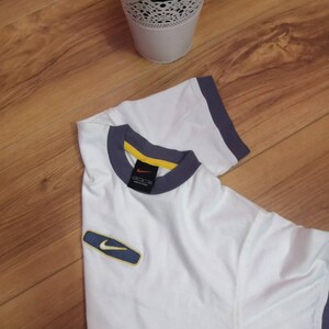 Peut inclure: T-shirt blanc avec bordure grise et jaune au col et aux manches. Un petit &eacute;cusson du logo Nike est sur la manche gauche. Le t-shirt est pli&eacute; sur une surface en bois.