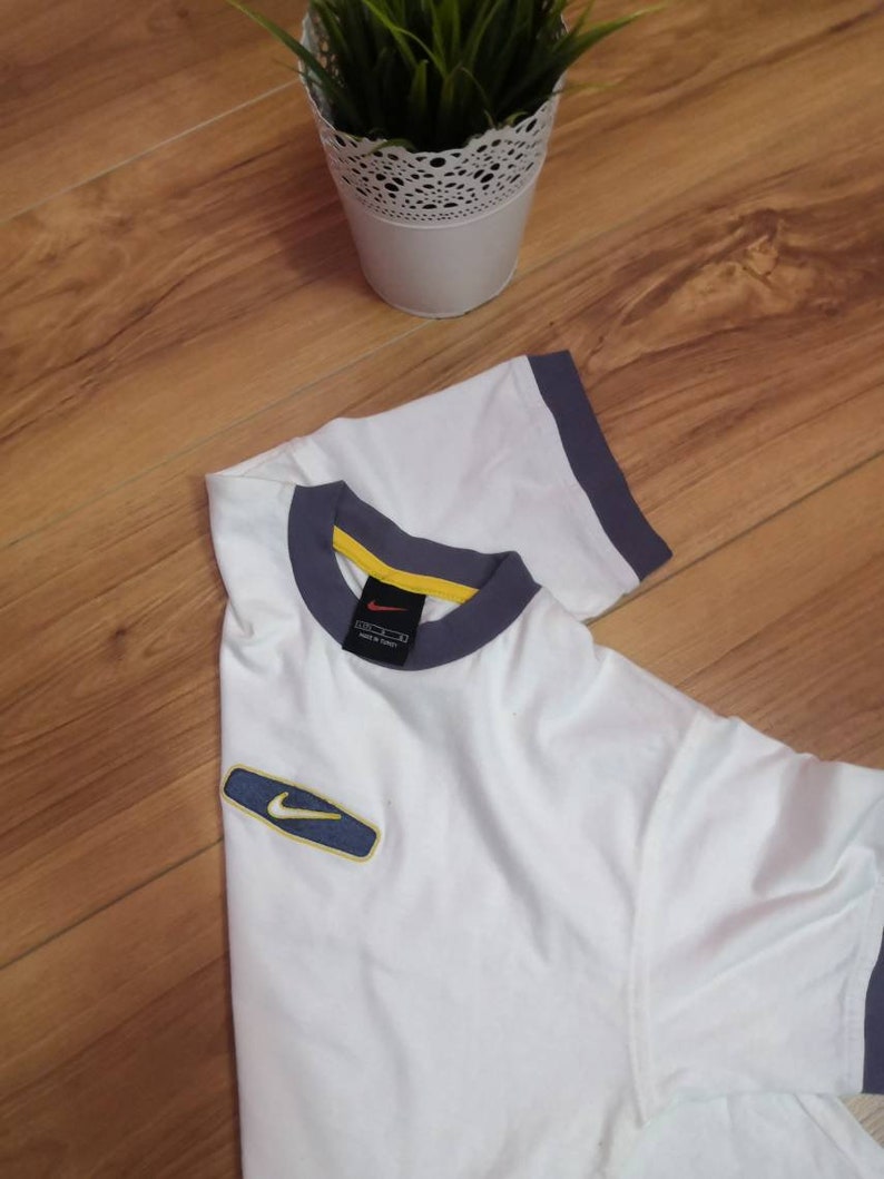 Peut inclure: T-shirt blanc avec bordure grise et jaune au col et aux manches. Un petit &eacute;cusson du logo Nike est sur la manche gauche. Le t-shirt est pli&eacute; sur une surface en bois.