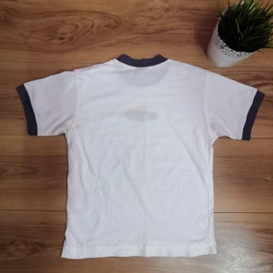 Peut inclure: T-shirt blanc avec bordure bleu marine au col et aux manches. Le t-shirt est pos&eacute; sur une surface en bois. Une petite plante en pot avec de l'herbe verte est en arri&egrave;re-plan.