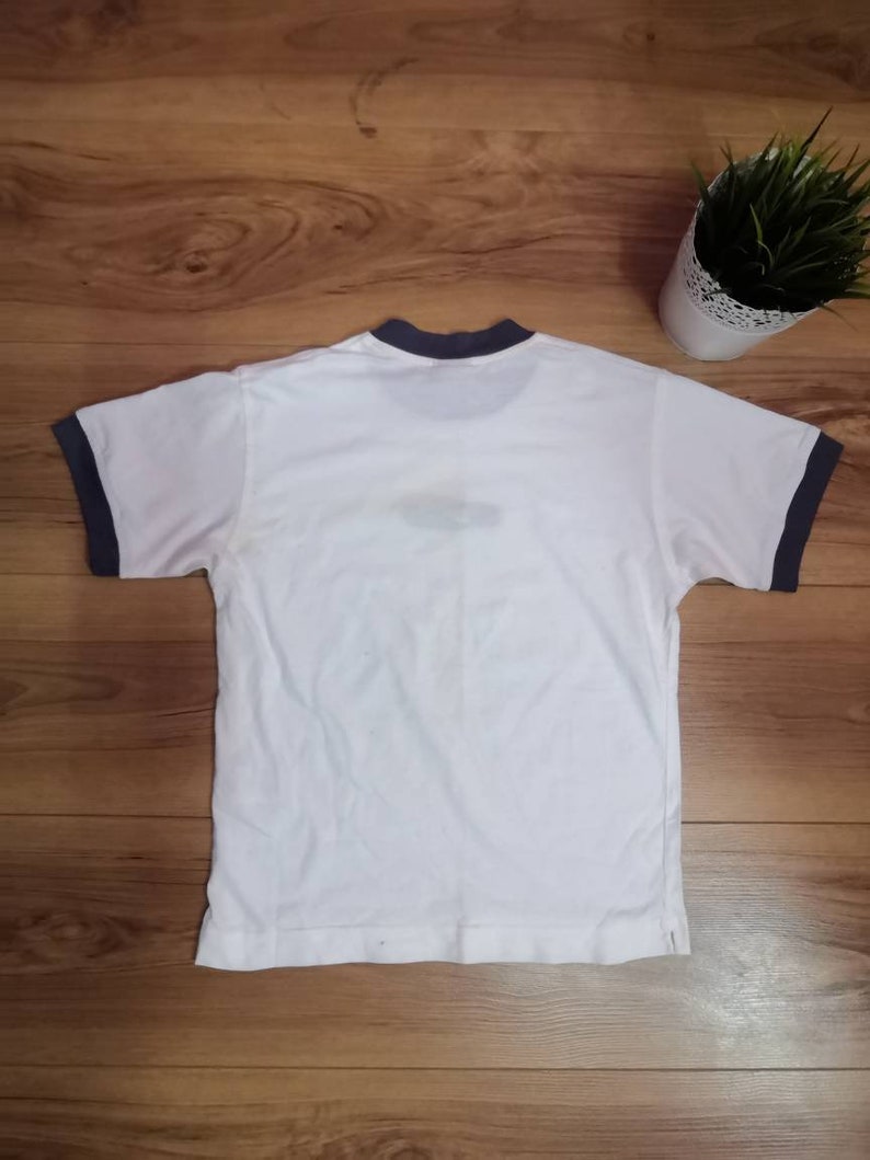 Peut inclure: T-shirt blanc avec bordure bleu marine au col et aux manches. Le t-shirt est pos&eacute; sur une surface en bois. Une petite plante en pot avec de l'herbe verte est en arri&egrave;re-plan.