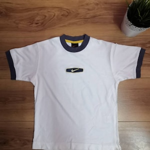 Peut inclure: T-shirt blanc avec bordure bleu marine et jaune. Le t-shirt pr&eacute;sente un petit logo rectangulaire avec un swoosh Nike. Il est &agrave; manches courtes et a un col rond.