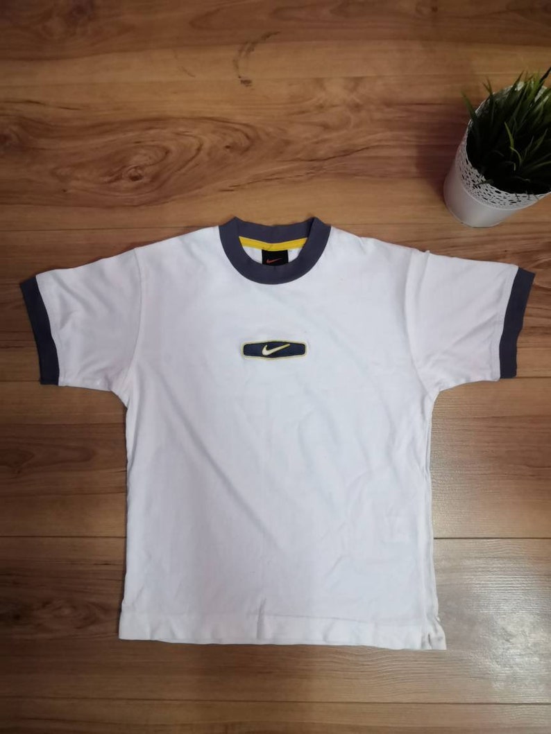 Peut inclure: T-shirt blanc avec bordure bleu marine et jaune. Le t-shirt pr&eacute;sente un petit logo rectangulaire avec un swoosh Nike. Il est &agrave; manches courtes et a un col rond.