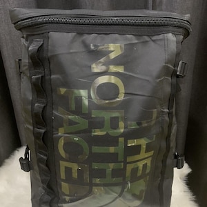 Op de afbeelding: Zwarte rugzak met de tekst "THE NORTH FACE" in een camouflagepatroon. De rugzak heeft een rechthoekige vorm, een ritssluiting aan de bovenkant en verstelbare bandjes. Hij heeft meerdere zijbanden en gespen.
