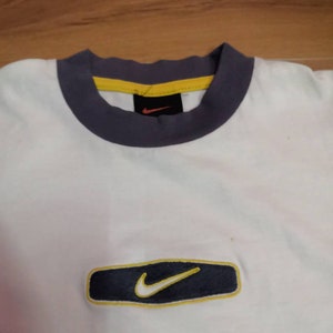 Peut inclure: T-shirt blanc avec une bordure bleu marine et jaune autour du col et un rectangle bleu marine avec une bordure jaune et un logo Nike swoosh blanc sur le devant. Le t-shirt est en mati&egrave;re souple.