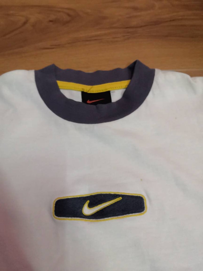 Peut inclure: T-shirt blanc avec une bordure bleu marine et jaune autour du col et un rectangle bleu marine avec une bordure jaune et un logo Nike swoosh blanc sur le devant. Le t-shirt est en mati&egrave;re souple.