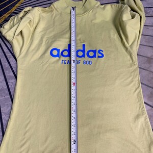 May include: A light olive green long-sleeve mock neck shirt with blue text reading adidas FEAR OF GOD. A vertical measuring tape is placed over the center, showing a length of approximately 27.5 inches from the collar to the bottom hem.