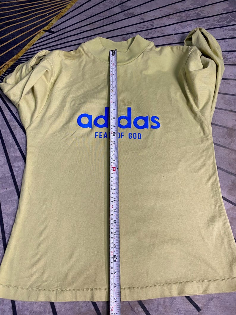 May include: A light olive green long-sleeve mock neck shirt with blue text reading adidas FEAR OF GOD. A vertical measuring tape is placed over the center, showing a length of approximately 27.5 inches from the collar to the bottom hem.