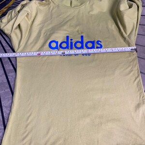 May include: A pale yellow long-sleeve mock neck shirt featuring the blue text "adidas" and "FEAR OF GOD" on the chest. A measuring tape is placed across the chest, showing a width of approximately 20 inches from side to side.