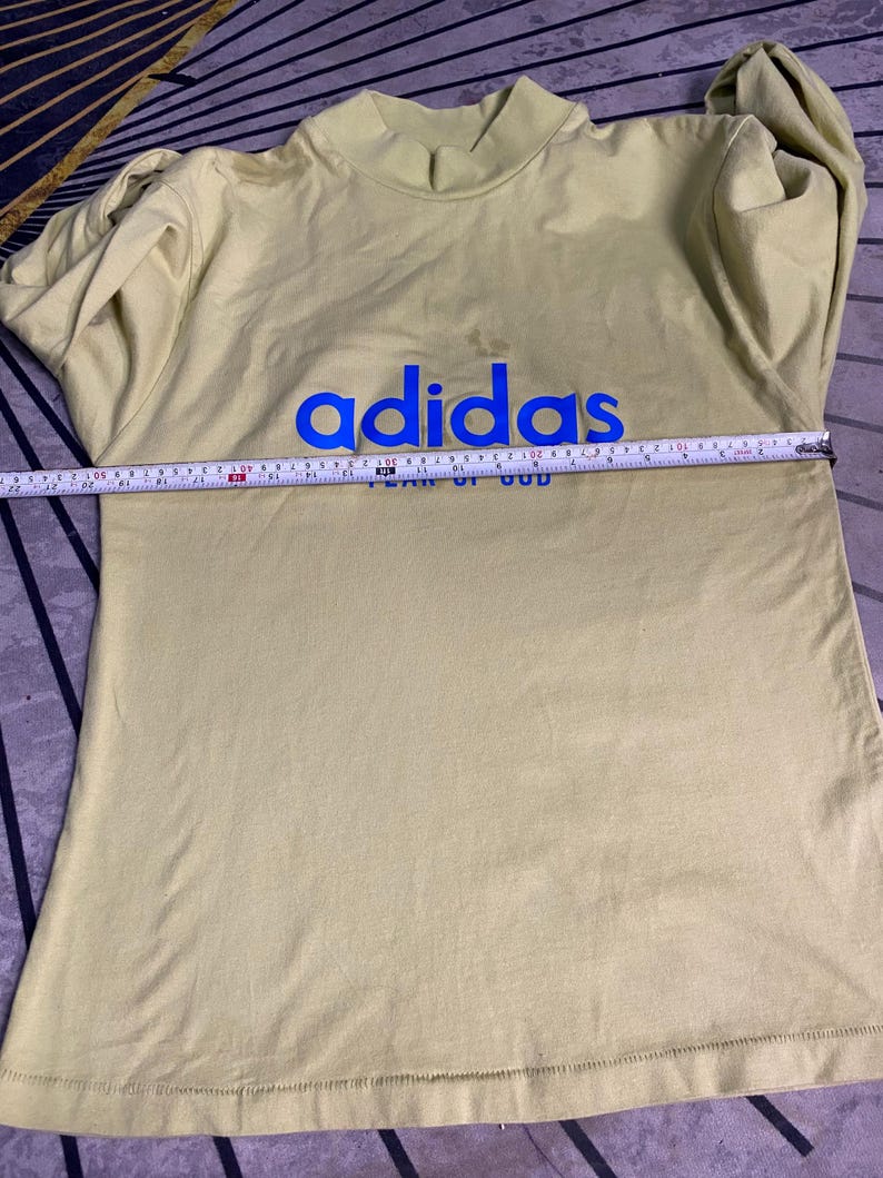 May include: A pale yellow long-sleeve mock neck shirt featuring the blue text "adidas" and "FEAR OF GOD" on the chest. A measuring tape is placed across the chest, showing a width of approximately 20 inches from side to side.