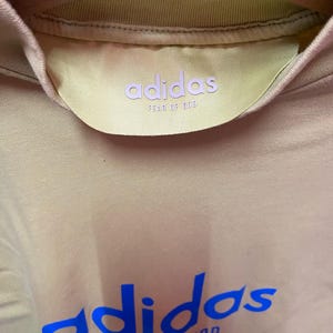May include: A beige t-shirt featuring a blue adidas and FEAR OF GOD logo printed on the chest. The interior collar displays a yellow satin-like neck label with the adidas and FEAR OF GOD branding in light purple text, hanging on a wooden hanger.