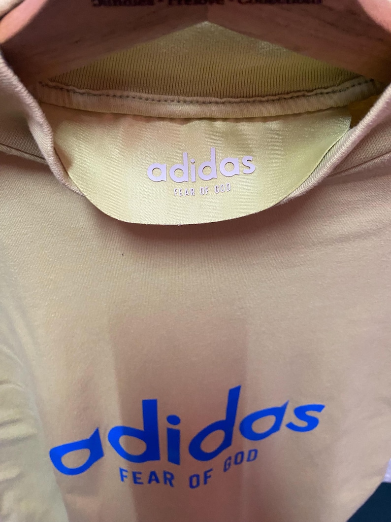 May include: A beige t-shirt featuring a blue adidas and FEAR OF GOD logo printed on the chest. The interior collar displays a yellow satin-like neck label with the adidas and FEAR OF GOD branding in light purple text, hanging on a wooden hanger.