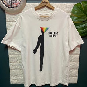May include: A white short-sleeve t-shirt featuring a black silhouette of a human figure with a rainbow-colored triangle at the head. The text GALLERY DEPT. is printed in black to the right of the figure. The shirt is displayed on a wooden hanger.