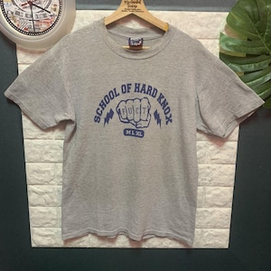 May include: A heather gray short-sleeve t-shirt with the text "SCHOOL OF HARD KNOX" in blue, along with a fist graphic. The shirt is hanging on a wooden hanger.