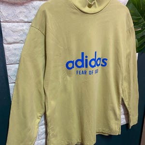 May include: A long-sleeve mock neck shirt in a muted olive green color. The front features the word adidas in blue lettering with the text FEAR OF GOD printed in smaller blue capital letters directly underneath the brand name.