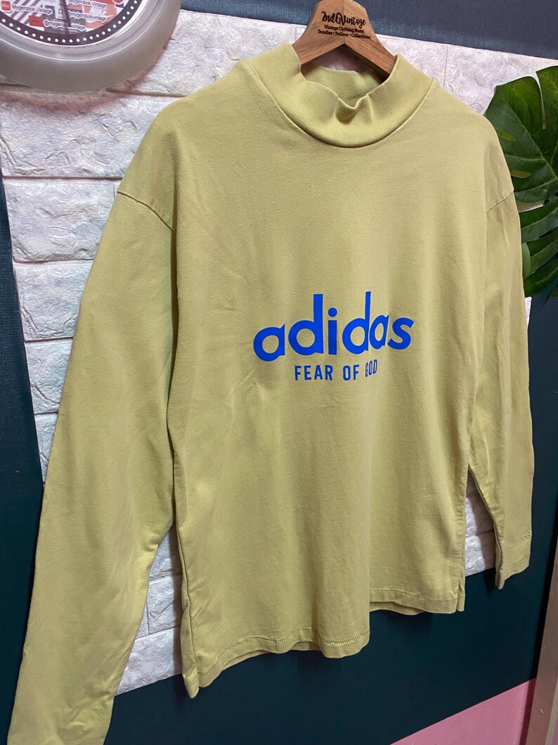 May include: A long-sleeve mock neck shirt in a muted olive green color. The front features the word adidas in blue lettering with the text FEAR OF GOD printed in smaller blue capital letters directly underneath the brand name.