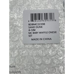 Pu&ograve; includere: Un'etichetta bianca con un codice a barre, codice prodotto BDBMC22398, colore DUNA DI SABBIA, taglia 6-12 mesi, descrizione SET DI BODY A GOFRE PER BAMBINI MC, e PRODOTTO IN CINA.