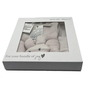 Pu&ograve; includere: Una scatola regalo bianca con una finestra trasparente che mostra un completo per neonato e un giocattolo di peluche. La scatola ha il testo "BAREFOOT DREAMS" e "For your bundle of joy" stampato su di essa.