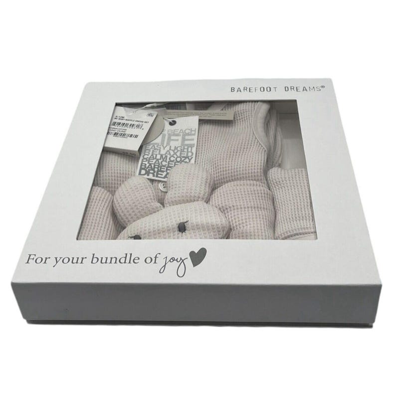 Pu&ograve; includere: Una scatola regalo bianca con una finestra trasparente che mostra un completo per neonato e un giocattolo di peluche. La scatola ha il testo "BAREFOOT DREAMS" e "For your bundle of joy" stampato su di essa.