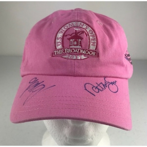 Op de afbeelding: Roze baseballpet met het US Women's Open logo en de tekst "The Broadmoor 2011". De pet is gesigneerd door twee mensen.