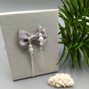 May include: A pair of silver earrings with a pearl and tassel design. The earrings are displayed on a gray box with a gray bow.