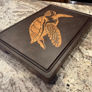 May include: A rectangular wooden cutting board with a dark brown finish. A light brown turtle design is inlaid on the surface. The board has a recessed juice groove around the perimeter.