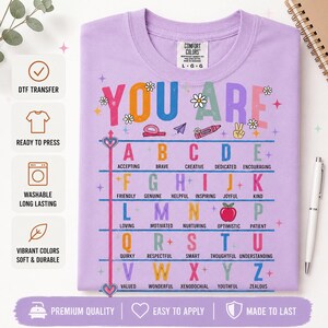 Puede incluir: Una camiseta de color morado claro con el texto YOU ARE en letras coloridas, seguido de una tabla alfab&eacute;tica donde cada letra corresponde a un adjetivo positivo. El dise&ntilde;o incluye peque&ntilde;os iconos como flores, un cray&oacute;n y un coraz&oacute;n, impreso en una camiseta de la marca Comfort Colors.