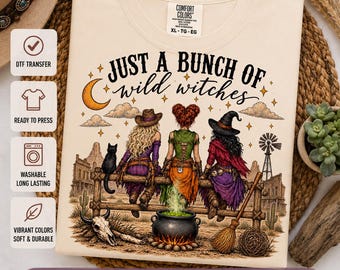 Puede incluir: Una camiseta de color crema con un gráfico de tres figuras sentadas en una valla de madera y el texto "JUST A BUNCH OF wild witches". El diseño incluye una luna creciente, un gato negro, un caldero burbujeante, una calavera y una escoba.