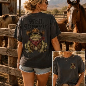 May include: A charcoal gray short-sleeve t-shirt featuring a graphic of a frog wearing a cowboy hat, vest, and red bandana. The back displays the text "Well, Sheeyit" above the frog, while the front features a smaller version of the same frog.